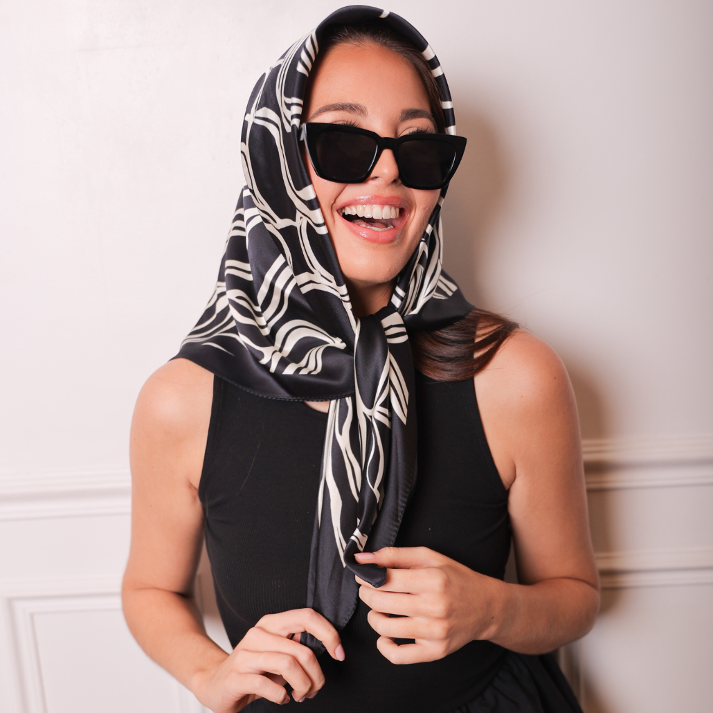 Femme qui porte un carré de soie sur la tête noir et blanc et qui sourit, elle porte des lunettes de soleil