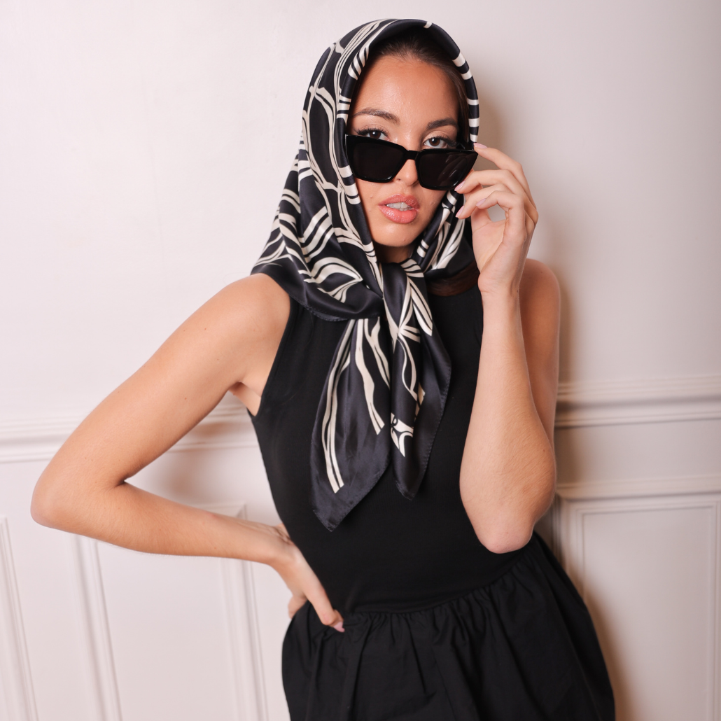 Femme qui porte un carré de soie noir et blanc avec une robe noire, elle porte de lunette de soleil légèrement baissés avec sa main
