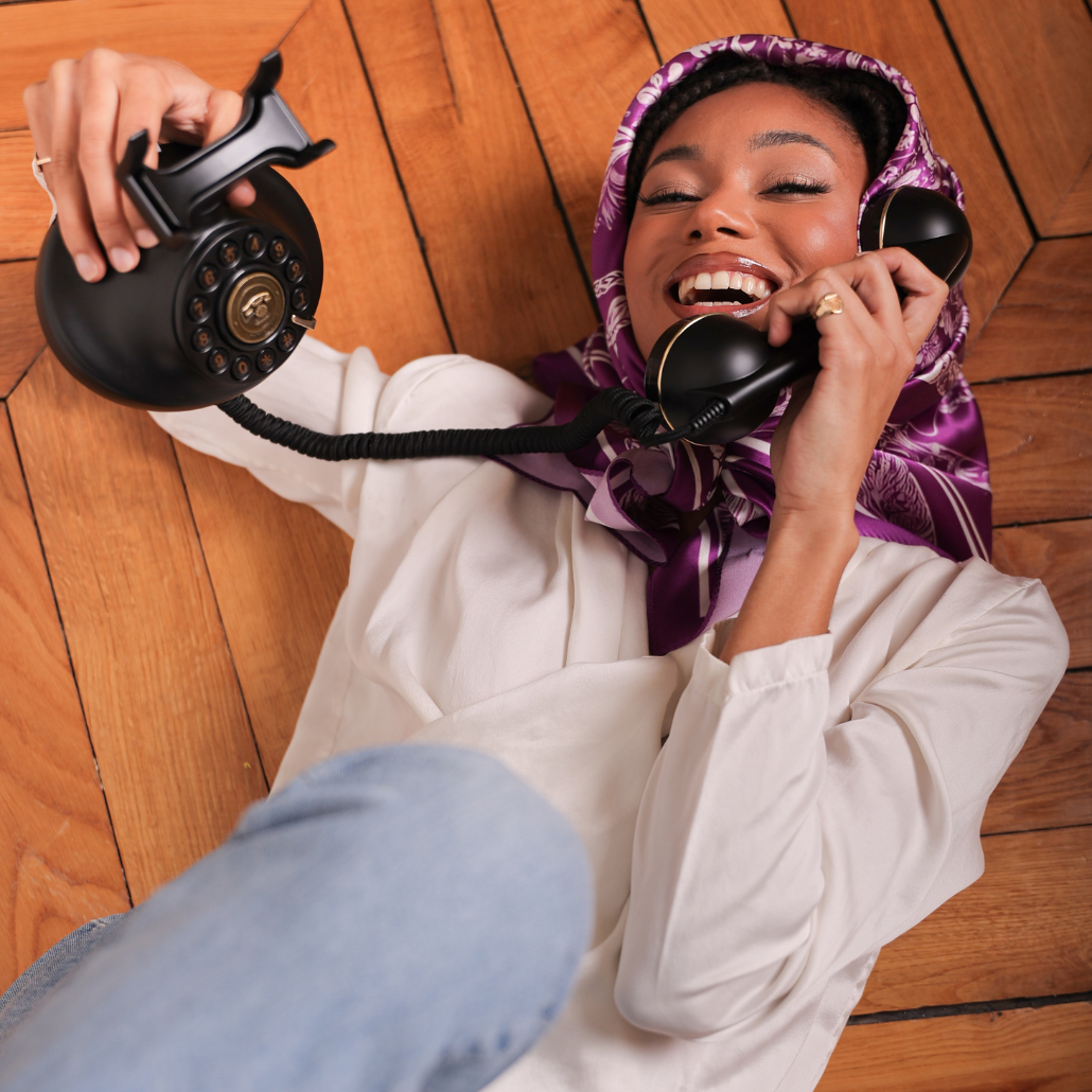 Carré de soie de couleur violet et blanc sur la tête qui est allongé sur un sol haussmanien et qui tiens à la main un téléphone vintage et qui sourit