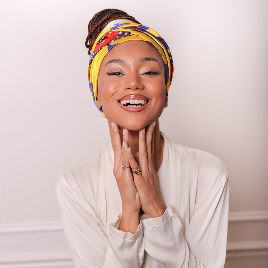 Femme qui porte un carré de soie imprimé wax couleur jaune, rouge et bleu, elle le porte en bandeau autour de ses braids attachées en chignon, elle affiche un beau sourire et porte une chemise blanche