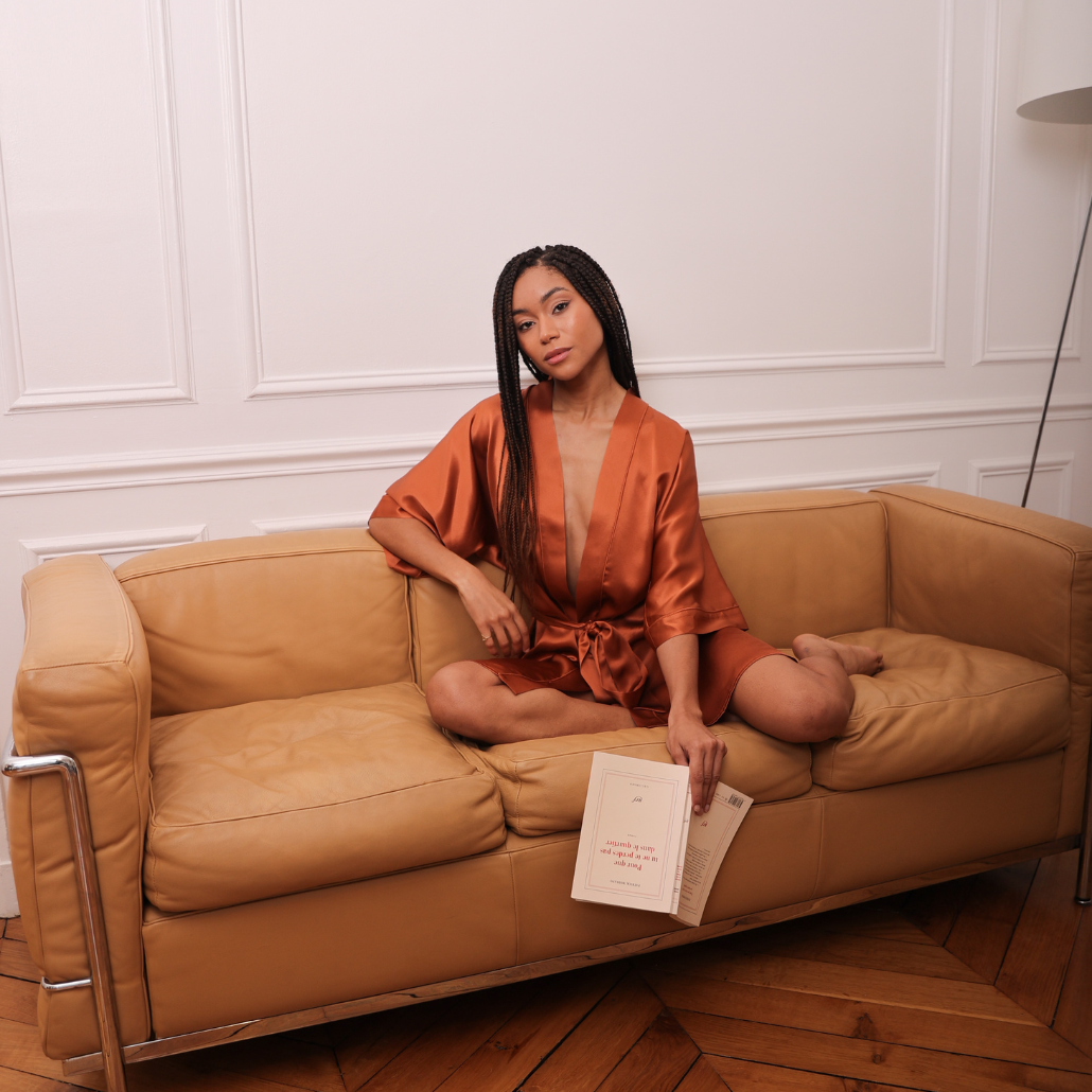 femme assise sur un canapé beige vêtue d'une robe de chambre terracota avec un livre à la main