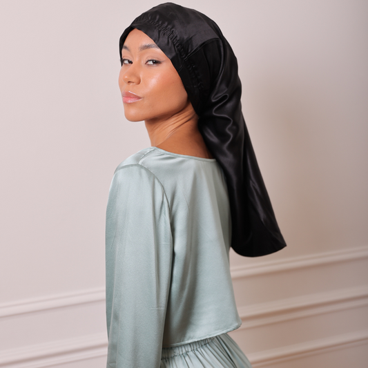 Femme qu pose de côté debout portant un pyamas couleur vert sauge et un bonnet de protection en soie long noir 