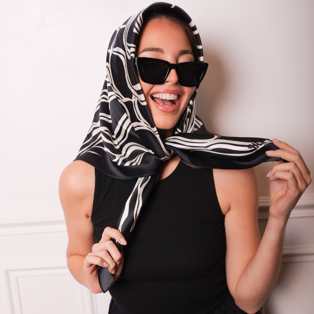 Femme qui porte un carré de soie noir et beige sur la tête, elle sourit avec des lunettes de soleil. 